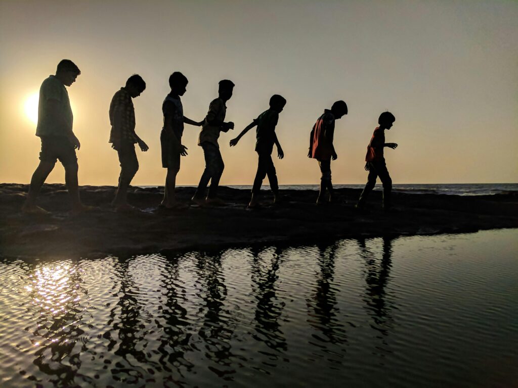 Home A group of children walk along the shoreline, silhouetted against a sunset, reflecting in the water.